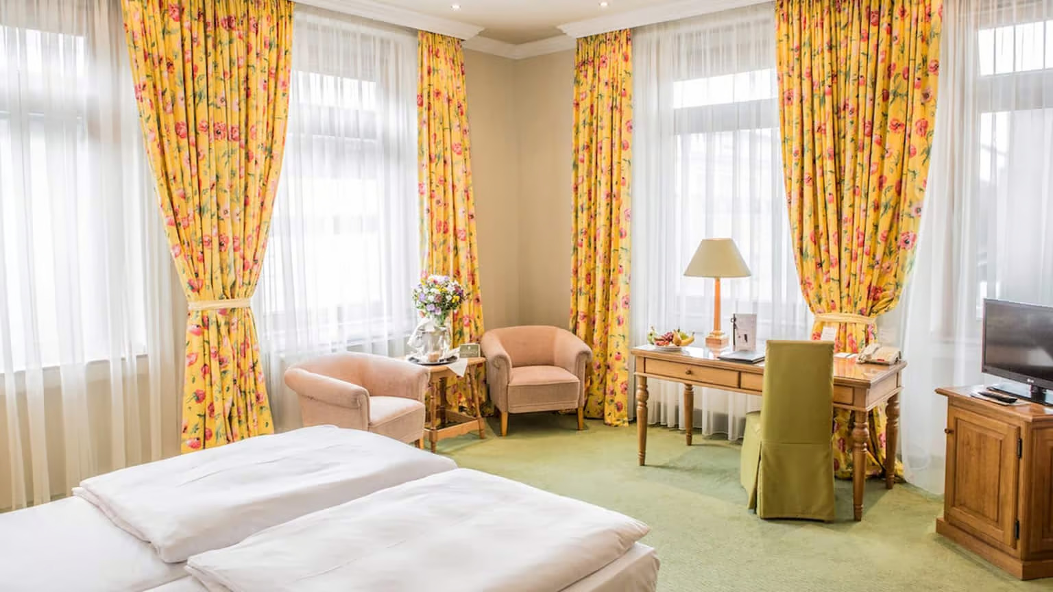 freundliches Doppelzimmer im Central-Hotel mit großem Bett, zwei Sesseln, gelb gemusterten Vorhängen, Schreibtisch mit grünem Stuhlbezug und Flachbildfernseher
